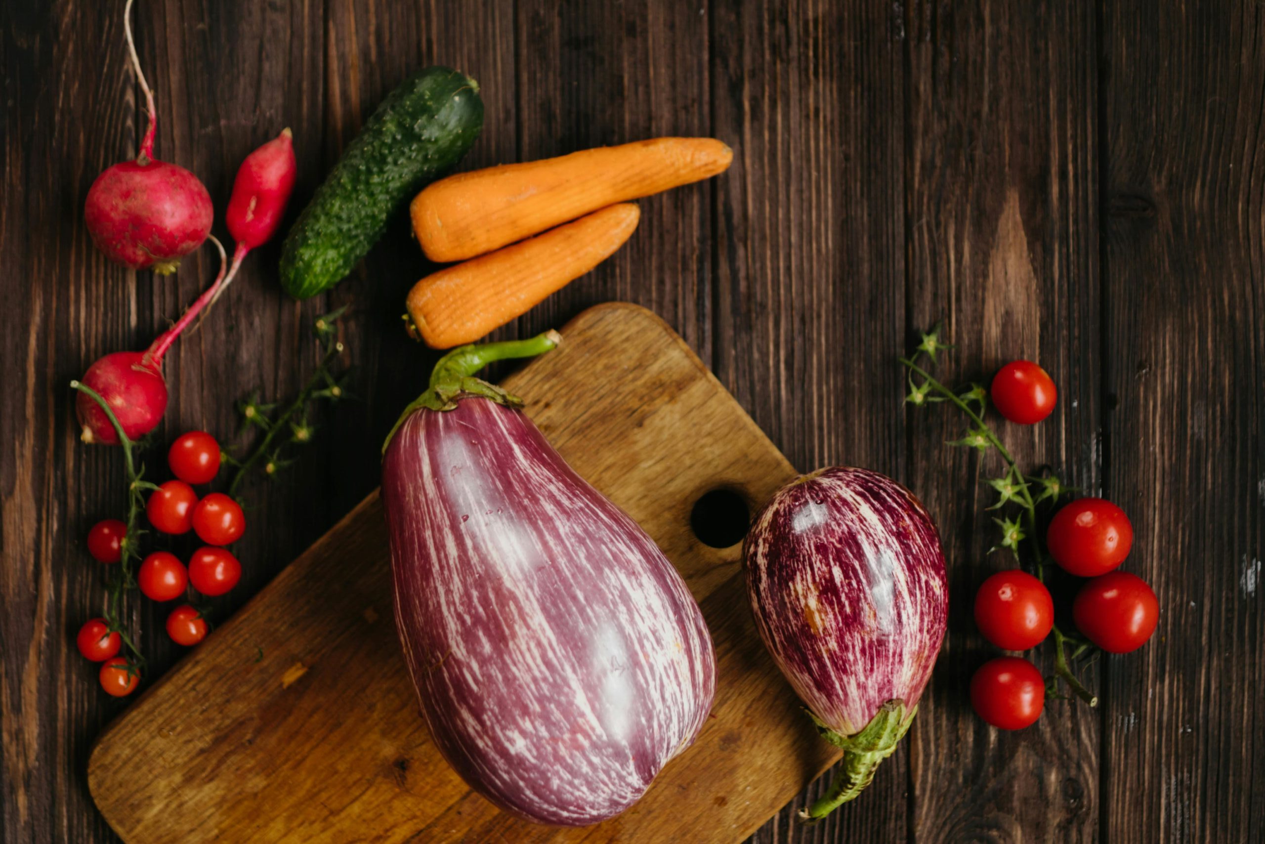 Top view of assorted fresh vegetables including carrots, eggplants, and cherry tomatoes on a wooden board.